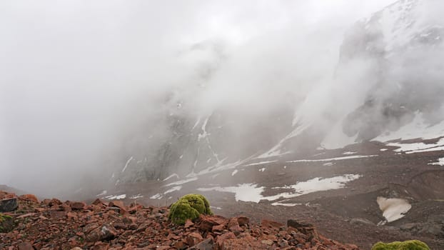 White clouds and snowy peaks. A lot of big stones. Steep cliffs, sometimes covered with white snow. Clouds float through the mountain tops. Moss grows in places. Mountains of the TRANS-ili Alatau.