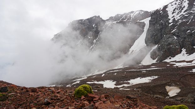 White clouds and snowy peaks. A lot of big stones. Steep cliffs, sometimes covered with white snow. Clouds float through the mountain tops. Moss grows in places. Mountains of the TRANS-ili Alatau.