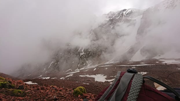 Backpack in the snowy mountains. Fog is coming. An Alpine camp in the rocky mountains, sometimes there is snow. Big clouds are passing. Mountain tops in the clouds. The ascent to the peak.