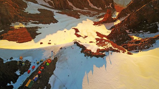 Mountaineering camp high in the snowy mountains. Epic red dawn, top view from a drone. Camp on the edge of the cliff. The snow-capped peaks, huge rocks. Preparing to climb the peak. Lots of tents.