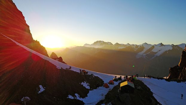 Mountaineering camp high in the snowy mountains. Epic red dawn, top view from a drone. Camp on the edge of the cliff. The snow-capped peaks, huge rocks. Preparing to climb the peak. Lots of tents.