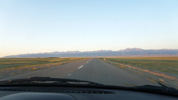 View through the windshield of the car on the road. The orange dawn is visible in the distance. The road is uneven, and the headlights illuminate the way. Oncoming cars are driving.