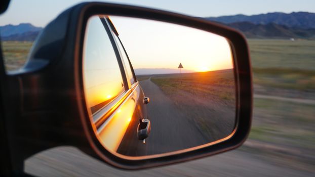 View in the side mirror of the car. Orange dawn over the hills. The car is going at high speed. Green fields, grass, and meadows are visible. Black color of the car.