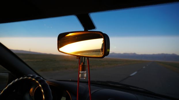 The rear view mirror of the car and dawn. Orange dawn over the hills. The car is going at high speed. Green fields, grass and meadows are visible. Black color of the car. Steering wheel, hood, glass.