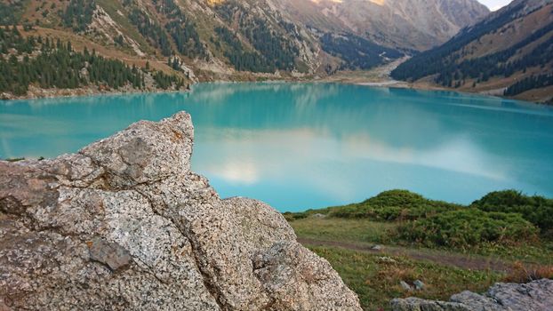 Big Almaty lake in the mountains. Green hills, flowers and fields. White clouds over the mountains. Mountain lake. In places there are stones. Protected place of nature, reserve of TRANS-ili Alatau