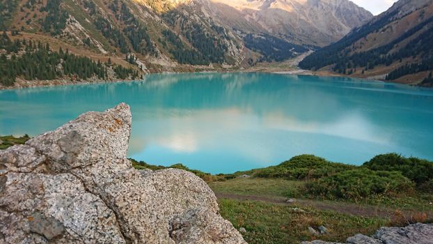 Big Almaty lake in the mountains. Green hills, flowers and fields. White clouds over the mountains. Mountain lake. In places there are stones. Protected place of nature, reserve of TRANS-ili Alatau