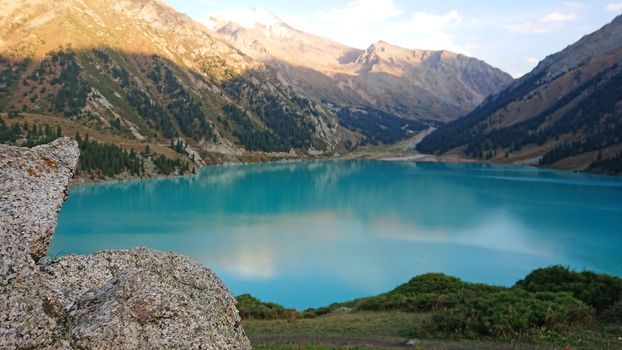 Big Almaty lake in the mountains. Green hills, flowers and fields. White clouds over the mountains. Mountain lake. In places there are stones. Protected place of nature, reserve of TRANS-ili Alatau