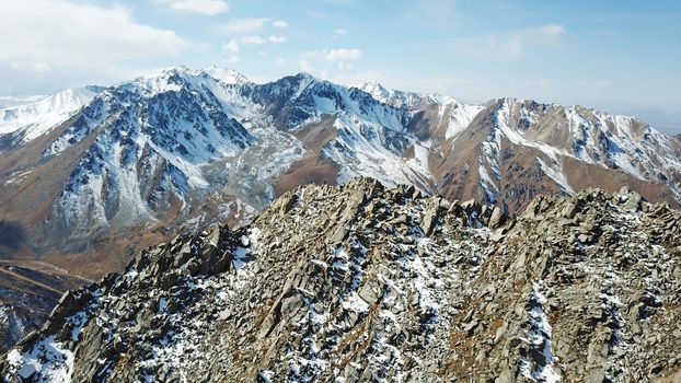 Top view of a group of tourists on top of a snowy peak. Huge rocks covered with snow. Climbers take photos from the top, pose. Flying above the mountains. Blue sky and steep slopes. Kazakhstan.