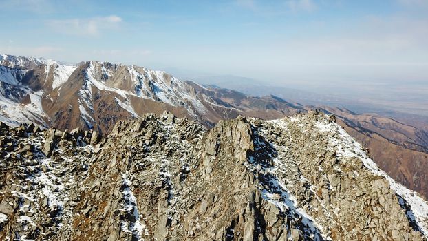 Top view of a group of tourists on top of a snowy peak. Huge rocks covered with snow. Climbers take photos from the top, pose. Flying above the mountains. Blue sky and steep slopes. Kazakhstan.