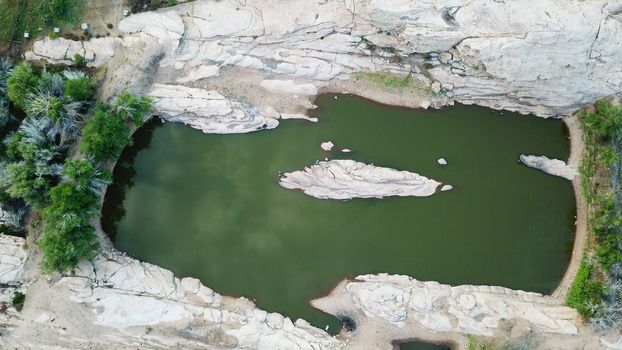 Green lake in the rocks. In the frozen lava. Green pit with an island in the middle. Grass and trees grow along the edges. Rocky terrain. Bektau-ATA Tract. Top view from a drone. Kazakhstan.