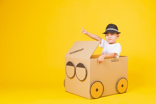 Portrait happy Asian cute little children boy smile so happy wearing white T-shirt driving car creative by cardboard and pointing finger, studio shot on yellow background with copy space