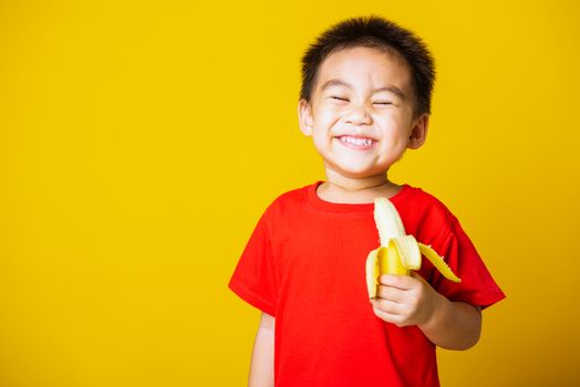 Happy portrait Asian child or kid cute little boy attractive smile wearing red t-shirt playing holds peeled banana for eating, studio shot isolated on yellow background