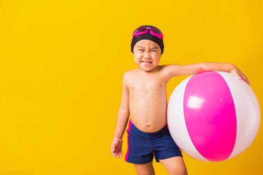 Summer vacation concept, Portrait Asian happy cute little child boy smiling in swimsuit hold beach ball, Kid having fun with inflatable ball in summer vacation, studio shot isolated yellow background