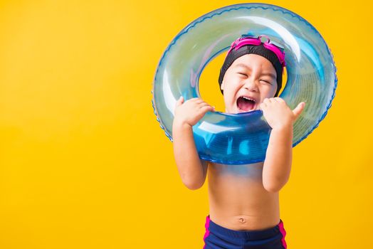 Summer vacation concept, Portrait Asian happy cute little child boy wear goggles and swimsuit hold blue inflatable ring, Kid having fun on summer vacation, studio shot isolated yellow background