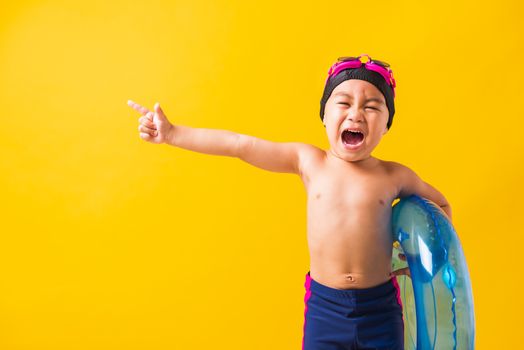 Summer vacation concept, Portrait Asian happy cute little child boy wear goggles and swimsuit hold blue inflatable ring, Kid hav fun point finger to side away, studio shot isolated yellow background