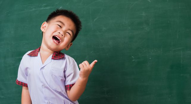 Back to School. Happy Asian funny cute little child boy from kindergarten in student uniform smiling point finger to side away space on green school blackboard, First time to school education concept