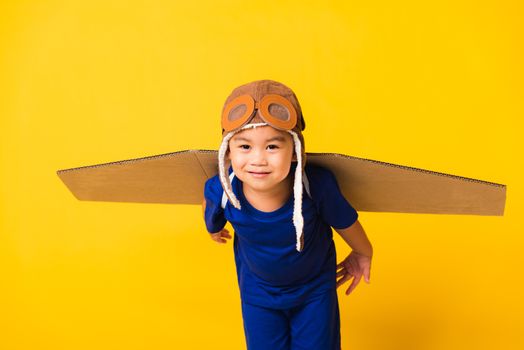Happy Asian handsome funny child or kid little boy smile wear pilot hat playing and goggles with toy cardboard airplane wings flying, studio shot isolated yellow background, Startup freedom concept