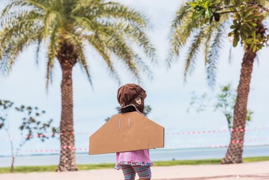 Happy Asian funny child or kid little girl smile wear pilot hat and goggles play toy cardboard airplane wing flying against summer sky cloud on trees garden background, Startup freedom concept