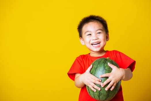 Happy portrait Asian child or kid cute little boy attractive smile wearing red t-shirt playing holds full watermelon that has not been cut, studio shot isolated on yellow background