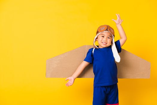 Happy Asian handsome funny child or kid little boy smile wear pilot hat play and goggles raise hand up with toy cardboard airplane wings flying, studio shot isolated yellow background, Startup freedom