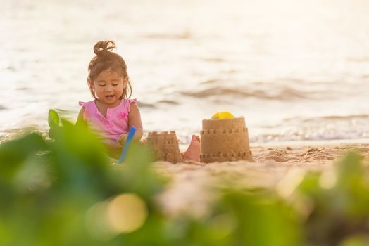 Happy fun Asian child cute little girl playing sand with toy sand tools at a tropical sea beach in holiday summer on sunset time, tourist trip concept