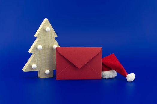 Red envelope for greetings next to a Christmas tree and Santa hat on a festive blue background. New Year and Christmas greeting season concept