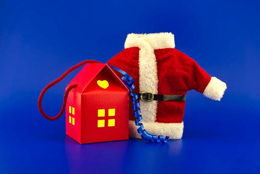 House shaped gift box with yellow glowing windows and red Santa Claus suit on a blue background. New Year and Christmas holiday season concept card decoration