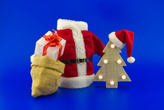 Red Santa Claus suit near stylized wooden Christmas tree with Santa hat and jute sack with gift boxes on a festive blue background. New Year and Christmas gift season concept