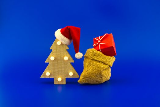 Stylized wooden Christmas tree with red Santa hat and jute sack with gift boxes on a festive blue background. New Year and Christmas gift season concept