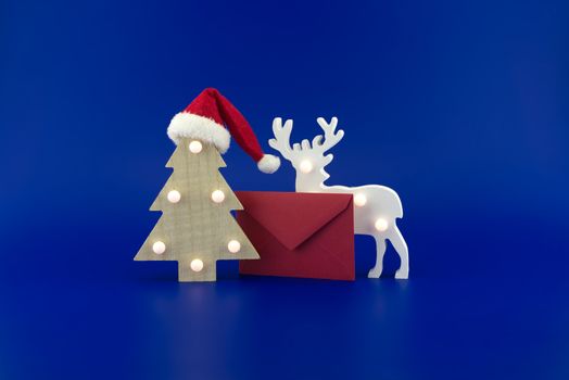 Christmas tree with a Santa hat on top next to a red envelope for greetings and stylized wooden reindeer on a festive blue background. New Year and Christmas greeting season concept