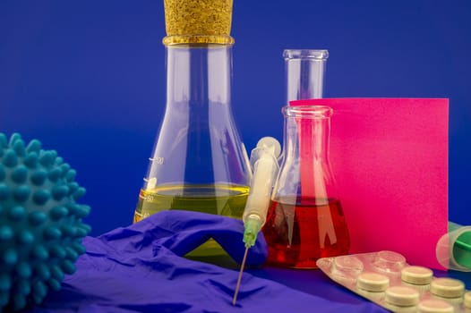 Concept of research for a virus vaccine with a conical lab flasks with yellow and red solutions alongside a hypodermic syringe and blue virus molecules with blank square of paper note for text