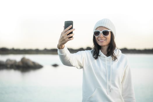 Young beautiful woman with big funny white sunglasses standing on a lake shore making a self portrait to sharing her winter vacation with her friends - Alone person doing selfie with smartphone