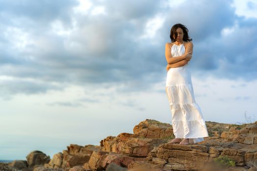 Caucasian woman wearing a long white dress with closed eyes standing on a rock with folded arms clutching herself in a sense of protection - Female person outdoor depressed for the solitude