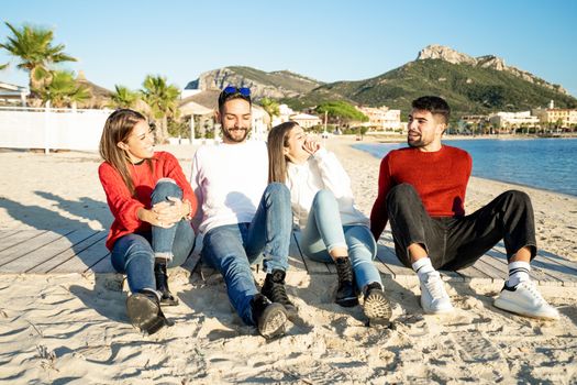 Two young couples by the sea at sunset in a seaside resort in winter having fun talking and laughing each other - Group of friends sitting on the beach in winter sea vacation - White and red clothing