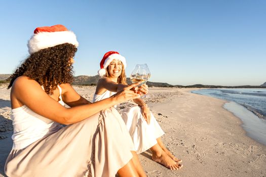 Lesbian couple with Santa hat sitting on seashore toasting with white wine glasses in winter vacation - Two beautiful happy women in love celebrating her Christmas and new year event on the beach