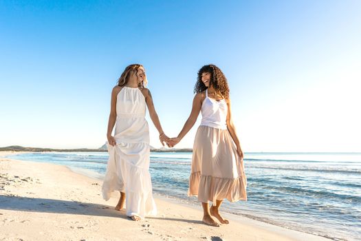 Tattoed beautiful blonde young Caucasian woman laughing holding the hand of her Black Hispanic girlfriend walking in the seashore in the morning or sunset - The beauty of different love between women