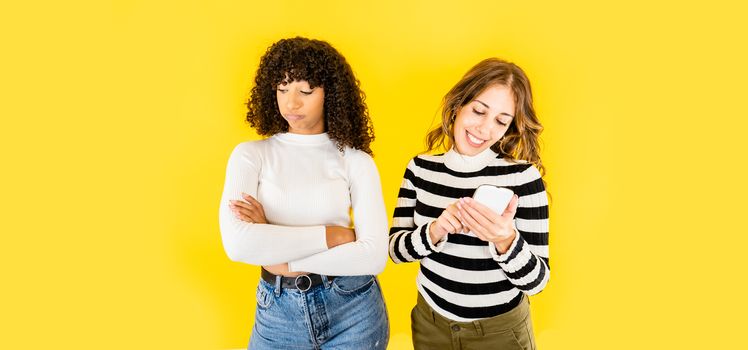 Two mixed race young women isolated funny picture on yellow background for copy space - Caucasian woman having fun with smartphone while her black Hispanic best friend snorts annoyed with folded arms