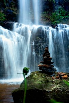Zen waterfall with lush foliage and balancing stones on a mossy green rock in foreground