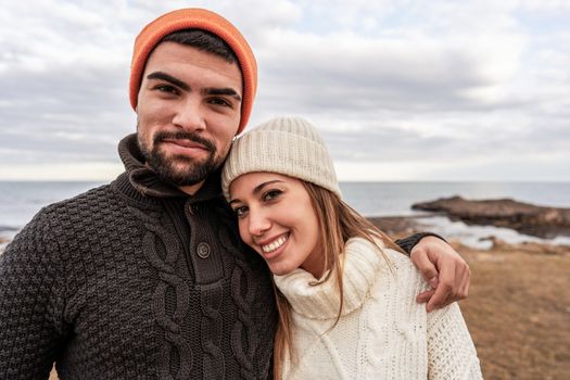 Beautiful young couple in love standing on sea resort looking at camera posing for portrait with cloudy sky in winter sea vacation - Caucasian girlfriend resting her head on shoulder of his boyfriend