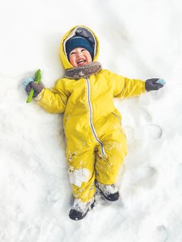 Smiling toddler in bright yellow jumpsuit is lying on snow. Laughing child walking outdoors in snowy weather. Top view on happy kid in colorful overall suit. Winter season.