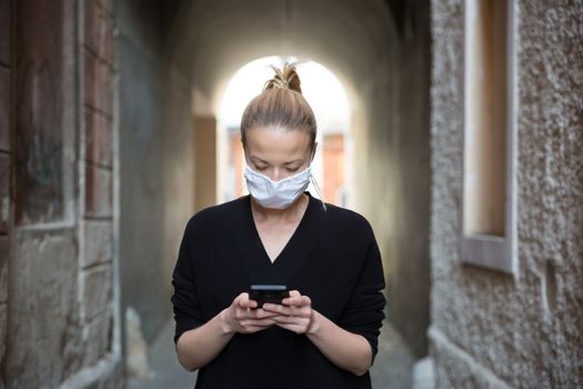 COVID-19 pandemic coronavirus. Casual caucasian woman at medieval city street using mobile phone, wearing protective face mask against spreading of coronavirus and disease transmission.