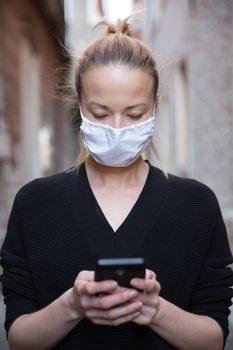 COVID-19 pandemic coronavirus. Casual caucasian woman at medieval city street using mobile phone, wearing protective face mask against spreading of coronavirus and disease transmission.