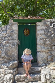 Beautiful young female tourist woman wearing big straw hat using moile phone, sitting in front of white vinatage wooden door and textured stone wall at old Mediterranean town.