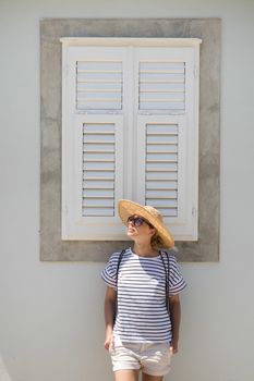 Beautiful young female tourist woman wearing sun hat, standing and relaxing in front of vinatage wooden window in old Mediterranean town while sightseeing on hot summer day.