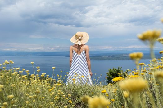 Rear view of young woman wearing striped summer dress and straw hat standing in super bloom of wildflowers, relaxing while enjoing beautiful view of Adriatic sea nature, Croatia.