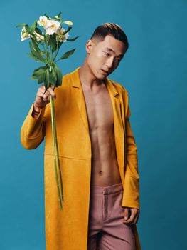 Charming Asian man with a bouquet of flowers and a yellow coat pink trousers nude torso blue background. High quality photo