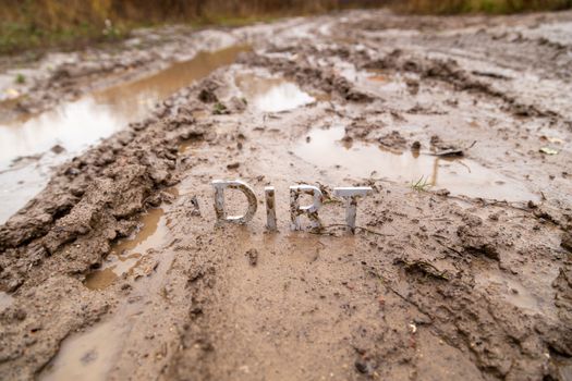 the word dirt composed of silver metal letters on wet clay surface with selective focus, background blur and linear perspective in cloudy autumn day light