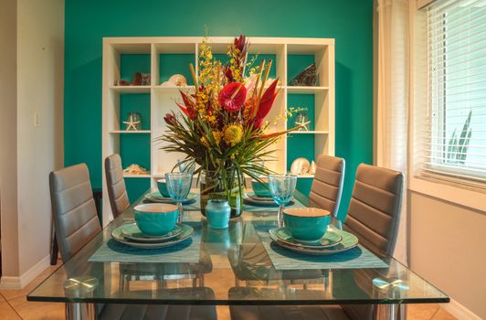 Tropical blue table set with flowers including Heliconia bihai, yellow oncidium orchids, yellow pincushion protea, and red anthuriums.