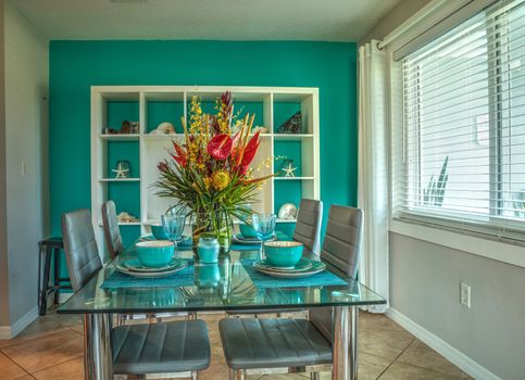 Tropical blue table set with flowers including Heliconia bihai, yellow oncidium orchids, yellow pincushion protea, and red anthuriums.