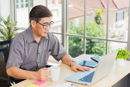 Asian executive senior businessman sitting on desk office with his office, the confident middle aged handsome man using laptop computer at workplace home office and drinking a cup of coffee